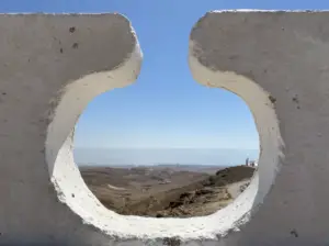השטח הסלעי והעקר, הנראה דרך חתך עגול בקיר לבן, משתרע מתחת לשמים כחולים צלולים. הסצנה הזו יכולה בקלות להיות חלק מכל בלוג טיולים בארץ, ללכוד את השלווה והיופי העז שהופכים טיולים בצפון הארץ לכל כך מושכים.