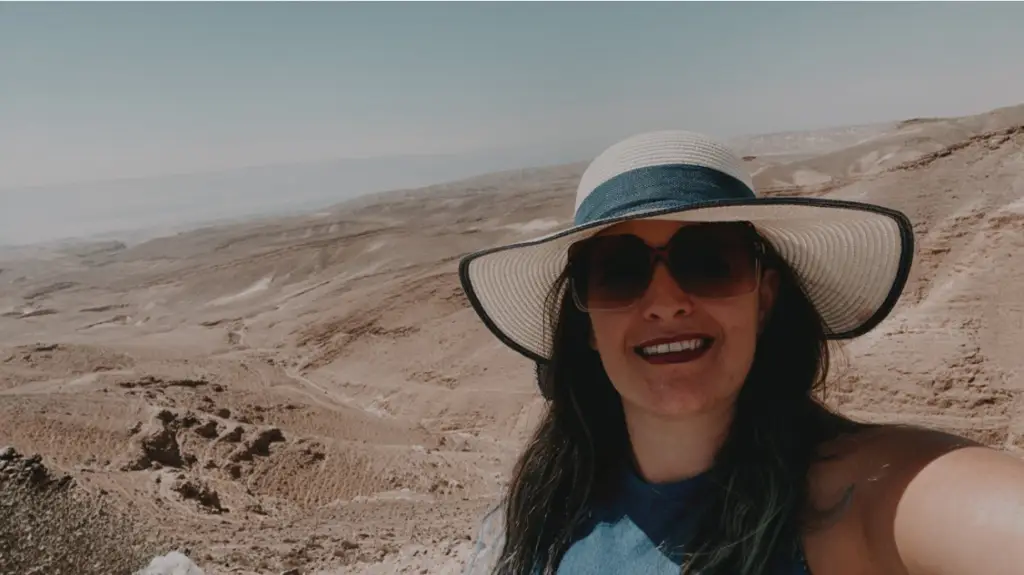 אישה עם משקפי שמש וכובע רחב שוליים מחייכת לסלפי בנוף המדברי, לוכדת את המהות של מטיילים בישראל. הרקע כולל גבעות חוליות מתגלגלות תחת שמיים כחולים צלולים.