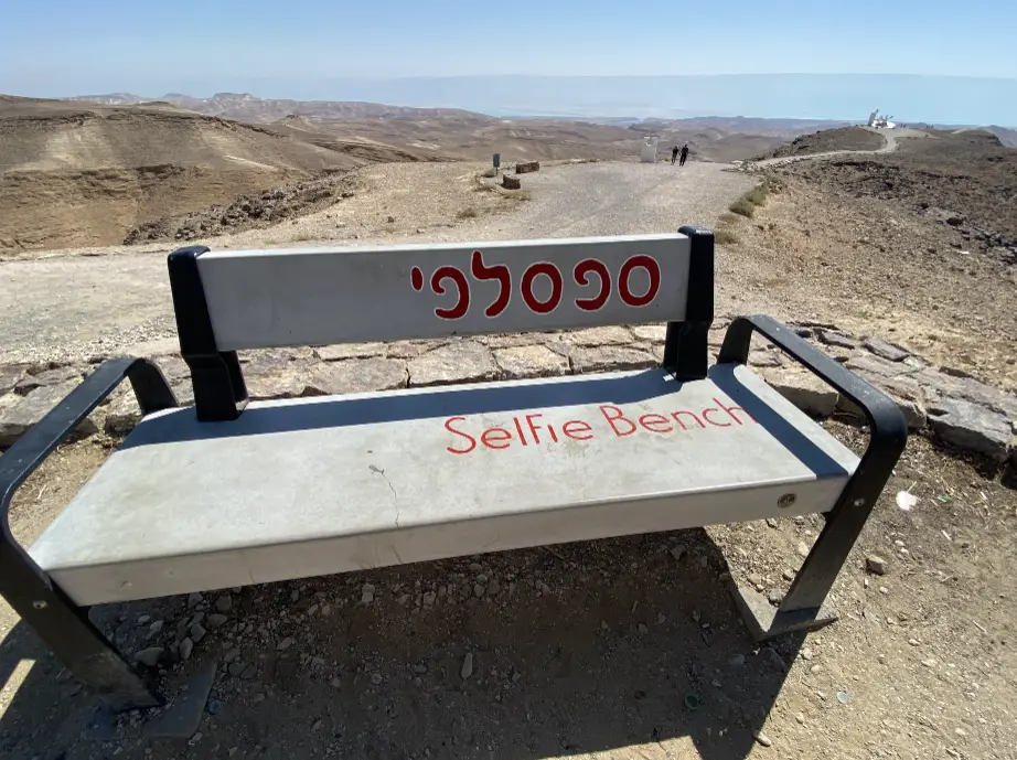 ספסל שכותרתו Selfie Bench באנגלית ובשפה נוספת יושב על נוף סלעי עם גבעות מרוחקות, לוכד את המהות של בלוג טיולים בארץ. שביל עפר מוביל משם, ומטיילים בישראל נראים חוקרים תחת שמיים כחולים צלולים.