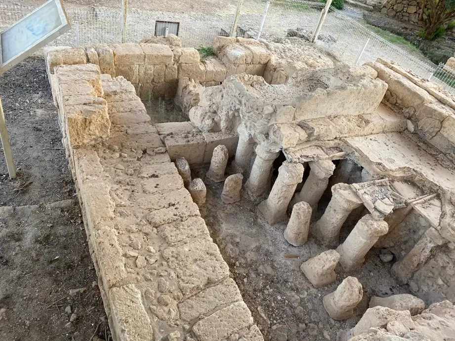 חורבות עתיקות עם קירות אבן ועמודים מעוותים, חפורות בחלקן, מוקפות בגדר נמוכה. משמאל נראה לוח. הקרקע הלא אחידה, מכוסה בעפר ופסולת אבן, מרמזת על היסטוריה. מושלם למטיילים בישראל או לטיולים בצפון הארץ בין צמחייה עבותה ברקע.