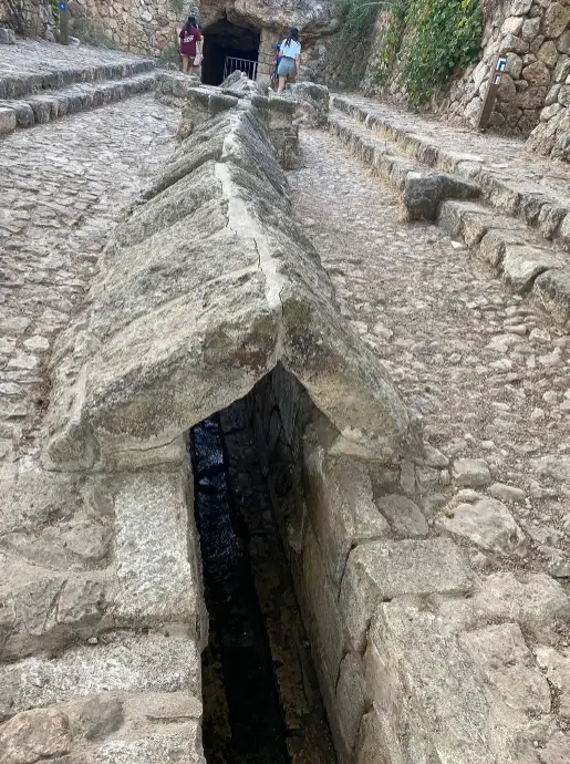 אמת מים עתיקה זו, השוכנת בתוך שביל צר מרוצף אבן, מתהדרת בתעלת מים מקורה העוברת במרכזה. שני אנשים מטיילים מרחוק, מוקפים בצמחייה עבותה הנצמדת לקירות האבן - אתר חובה למטיילים בישראל המחפשים אטרקציות ייחודיות.