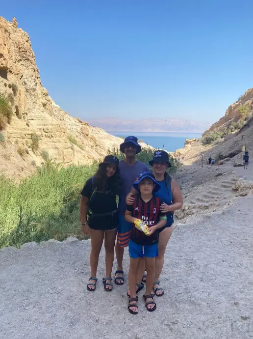 משפחה בת ארבע נפשות עומדת על שביל סלעי בנוף מדברי, מוקפת צוקים בצבע בז ושיחים ירוקים. לובשים כובעים ובגדים מזדמנים, הם נהנים מהנוף הנופי של גוף מים מנצנץ תחת שמיים כחולים צלולים - רגע מושלם לבלוג טיולים שלהם בארץ.