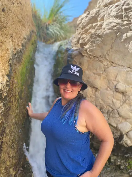 אדם שלובש גופיה כחולה, משקפי שמש וכובע דלי של אדידס מחייך כשהוא עומד ליד מפל קטן מוקף בתצורות סלע. עם יד אחת על המשטח הסלעי, הם מדיפים את הרוח של מטיילים בישראל.