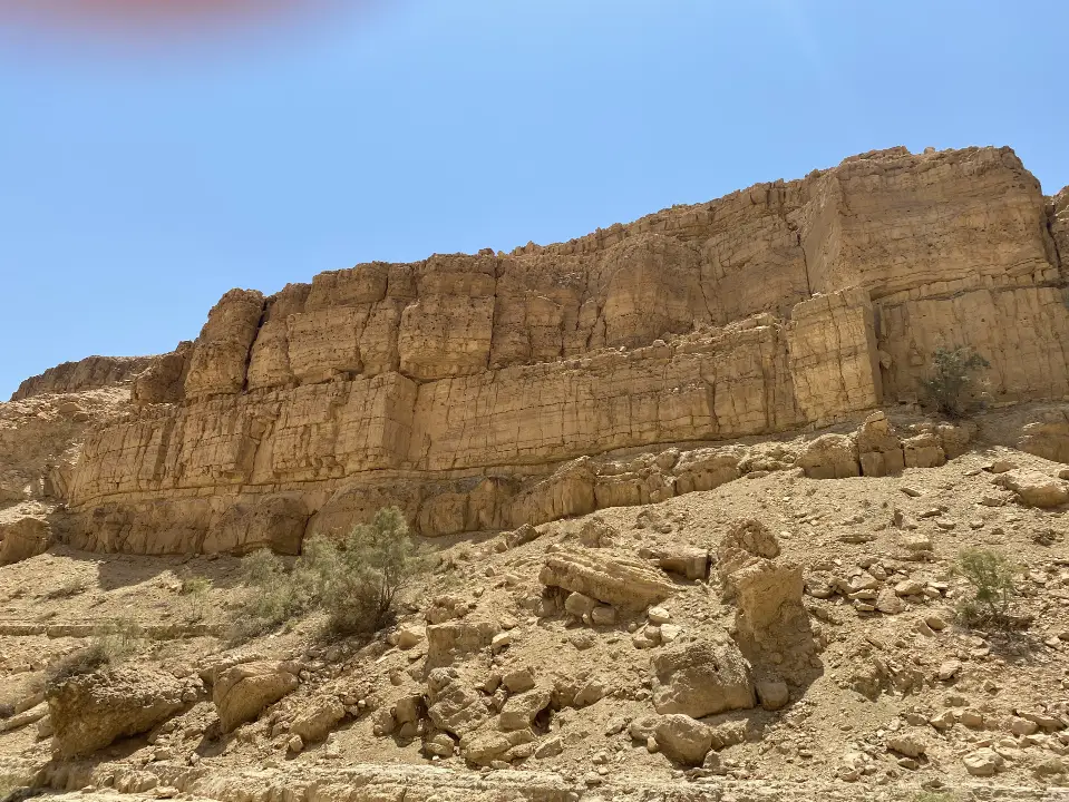 צוק מדברי מחוספס מתחת לשמים כחולים צלולים, הכולל שכבות של תצורות סלע חומות. בבסיסה ניתן לראות צמחייה דלילה, עם סלעים קטנים וסלעים מפוזרים. הנוף עוצר הנשימה הזה הוא אחד מני חוויות בטבע בישראל שאור השמש מטיל צללים חדים על פני המצוק.