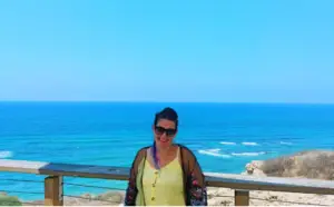 אישה במשקפי שמש וחלק עליון צהוב עומדת מחייכת מול מעקה עץ, לוכדת את המהות של חוויות בטבע בישראל. מאחוריה, הים הכחול והצלול והשמים הבהירים יוצרים תפאורה שטופת שמש ושלווה.