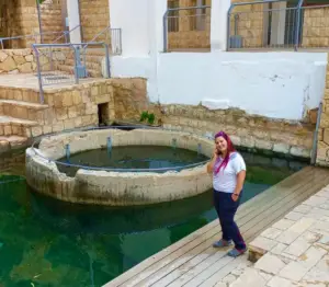 אדם עומד על במת עץ לצד מבנה בטון גדול ועגול מלא מים, מוקף חומות אבן ובניינים. מחייכים כשהם אוחזים בטלפון, הם תופסים את הרגע באחד ממקומות מומלצים לטיול בצפון הארץ.