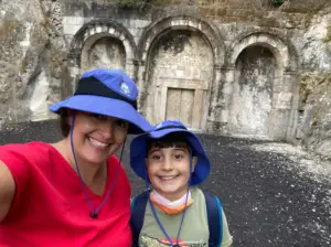 אישה וילד, שניהם חובשי כובעים כחולים, מחייכים למצלמה מול מבנה אבן בעל שלושה פתחים מקושתים. האדמה מכוסה בחצץ כהה, מה שמרמז על חוויה מרגשת של טיולים בצפון הארץ.