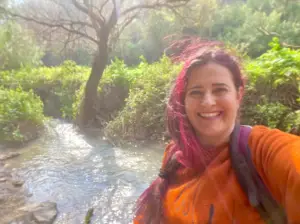 אדם עם שיער ורוד ארוך ותוסס מחייך תוך כדי פוזות ליד זרם קטן וזורם. הם לובשים טופ כתום ותיק גב באווירה טבעית שופעת וירוקה עם אור השמש המסנן בין העצים - רגע מושלם בטיולים בצפון הארץ.