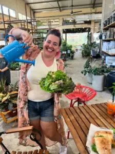 אישה המחזיקה קערת סלט גדולה מחייכת בעודה מוזגת בשובבות מים ממזלף כחול קטן בתוך הבית. לבושה בקימונו פרחוני, היא מביאה את הקסם של מטיילים בישראל פנימה, מוקפת בצמחים, שולחן עץ וכיסאות אדומים, לוכדת את המהות של חוויות בטבע בישראל.