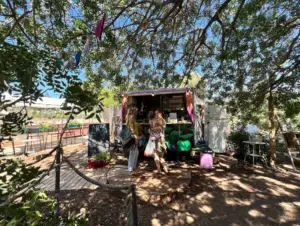 בית קפה קטן בחוץ בצל העצים מקבל את פני מטיילים בישראל. שני אנשים עומדים מלפנים, אחד מחזיק תינוק. דגלים צבעוניים ויצירות אמנות מלאות חיים מקשטים את ההתקנה. שולחנות וכיסאות פזורים על דק עץ ותופסים את חוויות בטבע בישראל ביום בהיר ושטוף שמש זה עם שמיים בהירים.