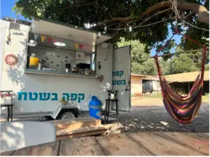 שוכן מתחת לעץ, בית הקפה המקסים של משאית האוכל מפתה מטיילים עייפים המטיילים במטיילים בישראל. בסמוך, ערסל צבעוני קורץ, ושולחן כפרי מזמין אתכם להירגע. ברקע מבנה עץ מוסיף לקסם של טיולים בצפון הארץ.