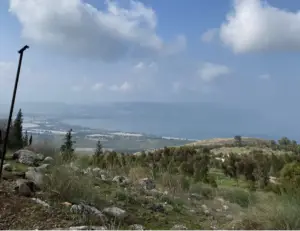 מבט על נוף הררי עם עצים וסלעים פזורים בחזית, המשקיף על גוף מים גדול תחת שמים מעוננים חלקית. קו חוף מרוחק ותפאורה הררית מפתים את מטיילים בישראל לחקור את היופי הטבעי ואת האטרקציות של האזור.