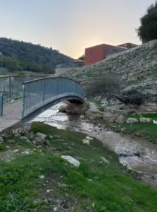 גשר להולכי רגל חוצה נחל קטן, מוקף בנוף עשב וסלעי. בניין אדום יושב על גבעה ברקע, עם עצים ושמים בהירים נראים. כשהשמש שוקעת, מטילה זוהר חם על הסצנה הציורית הזו, זה מושלם לשיתוף בבלוג שלך טיולים בארץ.