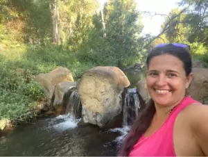 אישה בגופייה ורודה מחייכת למצלמה כשהיא עומדת מול מפל קטן עם נוף עם סלעים ועצים ירוקים, לוכדת את היופי של טיולים בצפון הארץ לבלוג טיולים בארץ.
