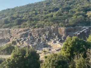 נוף מרהיב של צלע גבעה הכוללת תצורות אבן קשות בבסיסה, עם שמיכה עבותה של צמחייה ירוקה המכסה את המדרון. מושלם עבור אלה המחפשים חוויות בטבע בישראל, אור השמש מטיל צללים עדינים, מבליט את המרקמים של הסלעים והצמחים של האטרקציה הטבעית הזו.