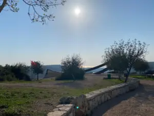 התמונה לוכדת סצנה חיצונית שטופת שמש מושלמת לטיולים בצפון הארץ, הכוללת עצים ושביל אבן. ברקע, פסל מתכת ניצב על רקע שמיים כחולים מעורפלים, כשהנוף נמתח לעבר גבעות רחוקות. דגל אדום מוסיף צבע ליד הפסל.