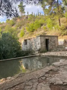 מבנה אבן קטן יושב ליד בריכת מים מלבנית, מוקפת נוף סלעי ועצים דלילים. בואו לחוות את היופי של טיולים בצפון הארץ כאשר צלע הגבעה ברקע מכוסה בשיחים ועצי אורן תחת שמיים בהירים.