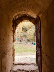 מציץ דרך קשת אבן עתיקה עם שער מתכת, הנוף מוביל לאזור גן עם עצים ואנשים מפוזרים, היוצרים תחושה של עומק והיסטוריה. הסצנה הציורית הזו היא אחד מהמקומות המומלצים לטיול המופיעים בבלוג טיולים בארץ.