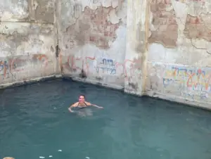 אדם שוחה בבריכה כפרית ומרובעת עם קירות בלויים ומכוסי גרפיטי מציע טוויסט לא צפוי על חוויות בטבע בישראל. המים השקטים מנוגדים את הקירות המסולפים, חושפים כתמים של לבנים חשופות וצבע דהה - פנינה שטרם התגלתה למטיילים בישראל.