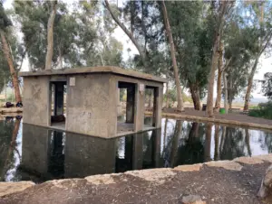 מבנה קטן מלבני מבטון שקוע בחלקו בבריכה שלווה מוקפת עצים גבוהים. האזור, המושלם למחפשי חוויות בטבע בישראל, משדר שלווה באווירה הכפרית הטבעית שלו וכולל שביל אבן המקיף את המים.