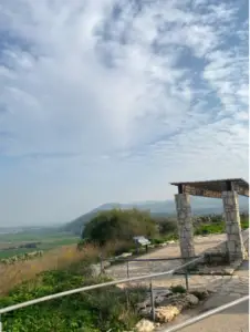 נוף מרהיב הכולל גזיבו מאבן עם גג עץ, על רקע גבעות מתגלגלות ושמים מעוננים חלקית. שביל עם מעקה מתכת מוביל אל הגזיבו, מוקף בצמחייה - מקום מושלם למטיילים בישראל המחפשים חוויות בטבע בישראל.