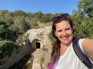 אישה עם שיער ארוך ומשקפי שמש לראשה מחייכת ומצלמת סלפי ליד מנהרת אבן מוקפת בירק ביום שמש. נהנית מחוויות בטבע בישראל, היא לובשת חולצה לבנה ונושאת תיק גב, חוקרת מקומות מומלצים לטיול בצפון.