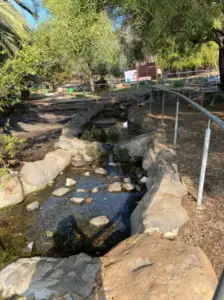 נחל צר זרוע אבנים מתפתל בפארק מוקף עצים, ומציע את אחת החוויות בטבע בישראל הקסומות. מעקה עוקב אחר שפת המים, וברקע נראה שלט עץ. אור השמש מסתנן בין העצים, מטיל צללים עדינים על הסצנה האידילית הזו מושלמת לטיולים בצפון הארץ.