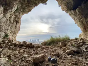 מתבונן מתוך פתח מערה סלעית, קו הרקיע של בניינים גבוהים משתרע למרחוק. בחזית, סלעים ממסגרים סיר בישול קטן. עננים מציפים את השמים כשהשמש מציצים מבעד, מושלם להרפתקה חוויות בטבע בישראל.