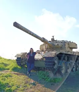 אישה עומדת מחייכת ליד מיכל ישן ובז על שדה עשב מתחת לשמים כחולים צלולים. הטנק החלוד מעט מרמז על ימים עברו, מושלם למטיילים בישראל שמחפשים מקומות ייחודיים. היא לובשת חולצה כחולה וגינס, נהנית מאחד ממקומות מומלצים לטיול הרבים.