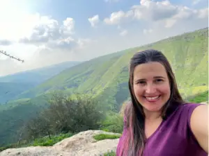 אישה חייכנית מצלמת סלפי מול צלע גבעה עבותה וירוקה עם רקע שמיים מעוננים באחד מאותם מקומות ציוריים מומלצים לטיול. היא לובשת חלק עליון סגול ועומדת על מדף סלעי, לוכדת את הנוף הרחב והנופי האופייני לטיולים בצפון הארץ.