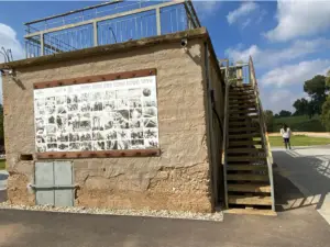 גרם מדרגות חיצוני מוביל לגג של מבנה אבן מלבני. קולאז גדול של תמונות בשחור-לבן מעטר את הצד, לוכד את הקסם של טיולים בצפון הארץ. ברקע, אדם הולך בשביל מרוצף מוקף דשא ועצים מתחת לשמים כחולים.