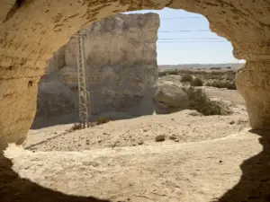 מבט מתוך מערה הצופה אל נוף מדברי סלעי. אור השמש מאיר את הקרקע החולית. מחוץ למערה נראים תצורת סלע ועמוד מתכת. צמחייה דלילה מפוזרת על פני השטח הצחיח, ומציעה הצצה להטיולים בצפון הארץ למטיילים בישראל המחפשים חוויות בטבע בישראל ייחודיות.