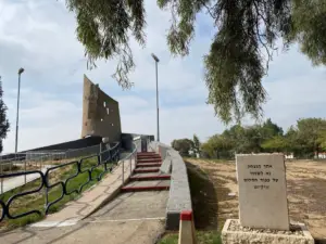 שביל המוביל אל אנדרטת אבן מודרנית ומופשטת מוקפת עצים ודשא, המציע סביבה שלווה לחוויות בטבע בישראל. שלט עם טקסט ממוקם בצד ימין בחזית תחת שמים מעוננים חלקית.