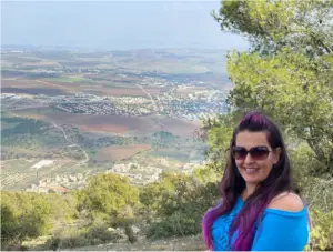 אישה עם משקפי שמש ושיער כהה ארוך עם הדגשות סגולות עומדת ליד עץ, מחייכת. היא לובשת חולצה כחולה. ברקע, האופייני לטיולים בצפון הארץ, נוף עצום עם שדות, מבנים פזורים ושמיים מעורפלים.