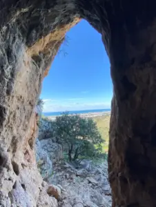מבט מתוך מערה סלעית הצופה אל נוף עם עצים ושדות מרוחקים מתחת לשמים כחולים. מושלם לטיולים בצפון הארץ, קירות המערה המרקמים ממסגרים את הנוף המדהים הזה, ממתינים להיחקר לקראת הבלוג הבא שלך בארץ.