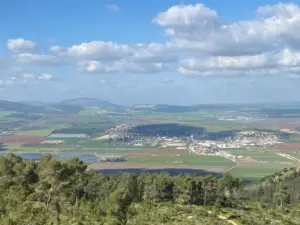 תצפית נוף מרהיבה בראש גבעה באחד המקומות מומלצים לטיול, עם שדות ירוקים ועיירה קטנה השוכנת בעמק. עננים מפוזרים מעטרים את השמים הכחולים מעל קו עצים בחזית, בעוד גבעות מתגלגלות יוצרות רקע עוצר נשימה.