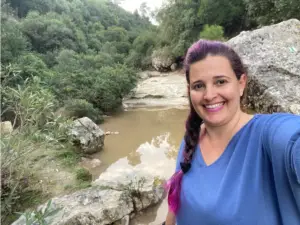 אדם עם צמה ושיער תוסס עומד מחייך מול נחל סלעי מוקף בצמחייה עבותה, ונהנה מאחד מהטיולים הרבים בצפון הארץ. הם לובשים חלק עליון כחול כשהמים זורמים בעדינות מאחוריהם, ממוסגרים על ידי עלווה וסלעים.