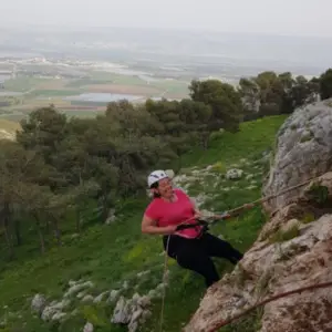 אדם עם קסדה וחולצה אדומה מטפס על סלעים במדרון עשב תלול, אטרקציה מלהיבה למחפשי הרפתקאות. באמצעות חבלים לתמיכה, הם מרחיבים את השטח על רקע של עצים שופעים ונוף סוחף - השראה מושלמת לבלוג טיולים בארץ.