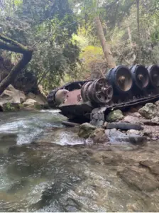 מיכל ישן וחלוד שוכב חלקית בתוך נחל צלול וסלעי מוקף יער עבות - פנינה נסתרת למטיילים בישראל. עצים וירק קשתים מעל המיכל, זורקים אור מנומר על המים, מה שהופך אותו לאחד ממקומות המומלצים לטיול לחובבי הטבע.