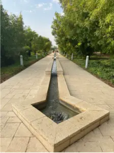 שביל אבן עם תעלת מים צרה עובר דרך פארק שלאורכו עצים ירוקים, ומציע את אחד ממקומות מומלצים לטיול. עמודי תאורה גבוהים נראים לאורך השביל, וכמה אנשים הולכים מרחוק תחת שמים מעוננים חלקית.
