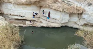 אנשים עומדים על מדף סלע ליד בריכת מים ירוקה ועכורה מוקפת צוקים סלעיים ועשב גבוה. אדם אחד שוחה בעוד אחרים מתכוננים להיכנס. נעליים וחפצים פזורים על המדף - סצנה מושלמת למטיילים בישראל המחפשים פנינים נסתרות בטבע.