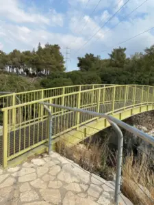 גשר צהוב עם מעקות מתכת משתרע על נקיק קטן, מוקף בצמחייה עבותה ועצים, מושלם למטיילים בישראל. השמיים מעוננים חלקית כשדרך אבן מובילה אל הגשר, כאשר קווי שירות מעליה מוסיפים לקסם של טיולים בצפון הארץ.