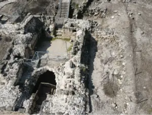 מראה אווירי של חורבות אבן עתיקות עם גרמי מדרגות וקירות בתוך שטח מחוספס וסלעי. מעקה מתכת מפאר גרם מדרגות, מעצים את גווני האדמה האפרפרים המגדירים את האדריכלות ההיסטורית הזו - מקום מושלם למטיילים בישראל לחקור אבני חן נסתרות.