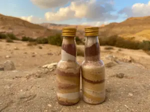 שני בקבוקי זכוכית מלאים בשכבות של חול ססגוני נחים על סלע, על רקע הנוף המדברי התוסס של צפון ישראל. התפאורה מציגה צמחייה פזורים וגבעות מתחת לשמים מעוננים, מושלם למטיילים בישראל המחפשים חוויות טבע ייחודיות.
