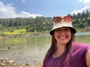 אישה חובשת כובע דלי פרחוני ומשקפי שמש מחייכת תוך כדי צילום סלפי על שפת אגם, אחד ממקומות רבים ומומלצים למטיילים בישראל. מאחוריה משתרעים עצים וצלע גבעה תחת שמים מעוננים חלקית.