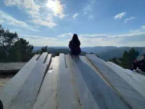 אדם עם שיער ארוך יושב על מבנה אבן גדול, מתבונן בנוף הררי תחת שמים מעוננים חלקית. עצים מקיפים את האזור ויוצרים תפאורה אידיליה למטיילים בישראל. אדם אחר יושב בקרבת מקום כאשר אור השמש משליך אפקט צללית שליו, מושלם לטיולים בבלוג בארץ.