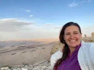 אישה מחייכת לסלפי בנוף סלעי, כשברקע הנוף המדברי העצום של ישראל תחת שמים מעוננים חלקית. היא לובשת קרדיגן לבן וחולצה סגולה, מצלמת חוויות בטבע בישראל בלתי נשכחות.