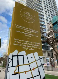 שלט גבוה לשביל העצמאות עם טקסט בעברית, ערבית ואנגלית מתאר מסלול הליכה בעשרה אתרים היסטוריים מ-1909 עד 1948. מפה בתחתית. בניין רב קומות ועצים ברקע ברחוב עירוני, מציע אטרקציות בתוך חקר עירוני בישראל.