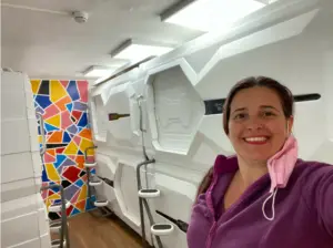 אישה מחייכת בחדר עם תרמילי שינה עתידני, מושלם לבלוג טיולים בארץ. היא אוחזת במסכה ורודה, ניצבת ליד ציור קיר גיאומטרי צבעוני. התרמילים כוללים ידיות וסולמות מתכתיים, מוארים בבהירות - יעד ייחודי לטיולים.