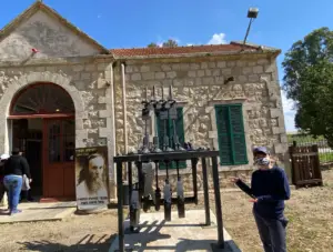 אדם עוטה מסכה מתייצב ליד פסל מתכת מול בניין אבן עם תריסים ירוקים, המציג את הקסם של מטיילים בישראל. אדם נוסף נכנס לבניין בו מוצגת ליד הכניסה כרזה עם דיוקן של גבר מבוגר.